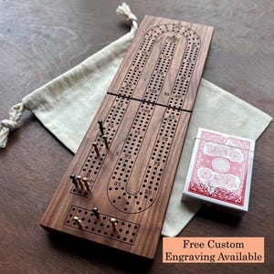 May include: A rectangular wooden cribbage board with a continuous track of drilled holes, featuring metal pegs in black and copper tones. A deck of playing cards with a red and white back design sits beside the board on a cream fabric drawstring bag.