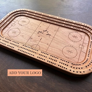 Peut inclure: Un plateau de cribbage en bois rectangulaire en forme de patinoire de hockey avec des coins arrondis. La surface présente des marquages de patinoire gravés, une feuille d'érable centrale et trois pistes de trous continues. Le texte ADD YOUR LOGO est inscrit.