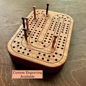 Peut inclure: Un plateau de cribbage rectangulaire en bois avec une piste en forme de U composée de trous percés. Le plateau comprend six pions métalliques aux finitions cuivre et noir. Une étiquette indique Custom Engraving Available.