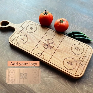 May include: Wooden hockey-themed chopping board with a handle, featuring engraved rink details and a hockey stick and puck emblem. Two red tomatoes and green peppers are placed on the board. The words "Add your logo" are visible.
