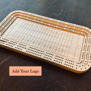 Peut inclure: Un jeu de cribbage en bois clair avec un motif de terrain de football gravé au centre. Le jeu comporte plusieurs rangées de trous pour les chevilles et le texte "Add Your Logo" sur un rectangle pêche.