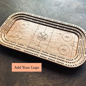 Peut inclure: Un jeu de cribbage en bois avec un design de patinoire de hockey. Le plateau est rectangulaire avec des coins arrondis et comporte des lignes et des cercles gravés. Le texte "Add Your Logo" est sur une étiquette rectangulaire.