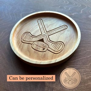 May include: A circular wooden coaster with a hockey design. The coaster has an engraved image of crossed hockey sticks and a puck. The coaster is made of light-coloured wood and has a smooth finish. The text "Can be personalized" is visible.