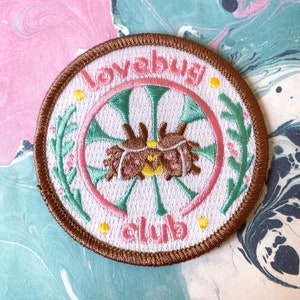 Op de afbeelding: Een bruin geborduurd patch met een witte achtergrond. De patch heeft een roze en groen bloemmotief met een bruin en gele bug in het midden. De tekst "Lovebug Club" is geborduurd in roze rond de rand van de patch.