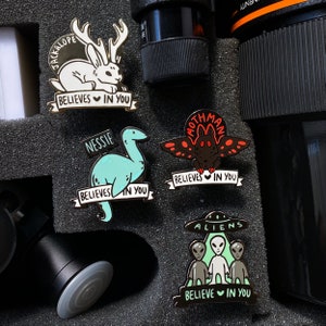 May include: Five enamel pins featuring different mythical creatures. The pins are gold and black with colorful accents. The pins say "Believe in You" and feature a jackalope, a mothman, a loch ness monster, and aliens.
