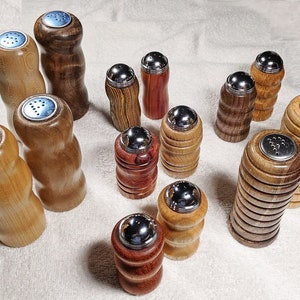 May include: A collection of 14 wooden salt and pepper shakers with silver tops. The shakers are made from different types of wood, including maple, cherry, and walnut. Some of the shakers have a smooth finish, while others have a more rustic look.