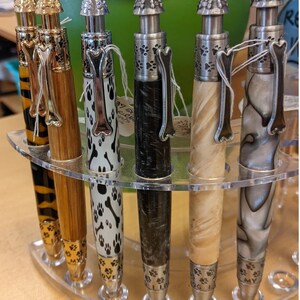 May include: A collection of dog-themed pens displayed in a clear acrylic holder. The pens feature various designs, including paw prints and bone-shaped clips. The text "I LOVE MY DOG" is stamped around the tip of each pen.
