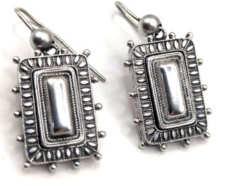 May include: A pair of silver-toned rectangular earrings. Each earring features a central, raised rectangle within a detailed frame. The frame is adorned with small, oval-shaped accents and tiny ball-shaped embellishments. The earrings have a hook closure.
