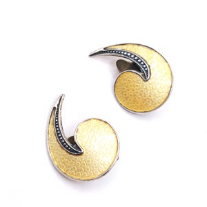 Peut inclure: Une paire de boucles d'oreilles dorées avec un motif semi-circulaire texturé. Chaque boucle d'oreille présente un accent argenté avec une ligne de petits points foncés. Les boucles d'oreilles sont posées sur un fond blanc.