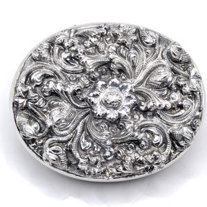 May include: Oval, silver-toned compact with intricate floral and foliate relief detailing. The design features a central flower surrounded by swirling leaves and blossoms, creating a textured, ornate surface. The compact has a polished, reflective finish.