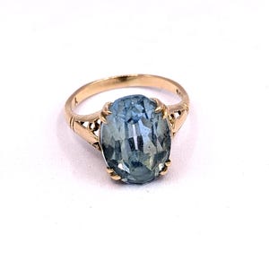 May include: A gold ring featuring a large, oval-cut, light blue gemstone. The ring's band has decorative filigree detailing on the sides. The gemstone is securely set with gold prongs, showcasing its clarity and color.