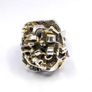 May include: A silver ring with an abstract design. The ring has a textured surface with a mix of silver and gold-toned metal. The rectangular face is decorated with geometric shapes. The ring's design is artistic and modern, suitable for everyday wear.