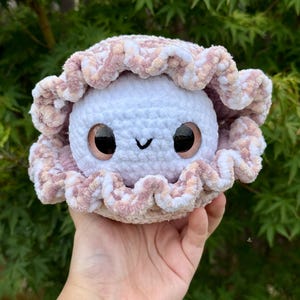 May include: A handmade crochet plushie in the shape of a clam with a smiling face. The plushie has a white body, large black eyes, and a pink and white ruffled shell. The plushie is held in a hand against a backdrop of green foliage.