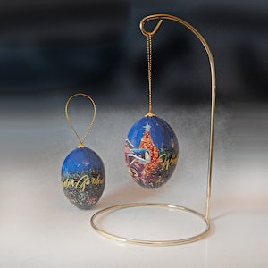 May include: Two blue Christmas ornaments with gold accents. One ornament is hanging from a gold stand, the other has a gold loop. The ornaments feature a winter scene with a Christmas tree and the words "Winter Garden".