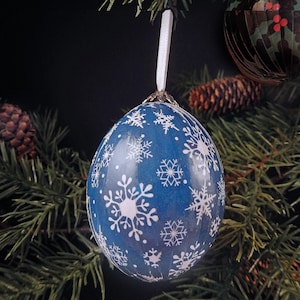 May include: A blue Christmas ornament with white snowflake designs hangs from a white ribbon. The ornament is round and has a metallic cap. The background includes pine needles, pine cones, and another ornament.