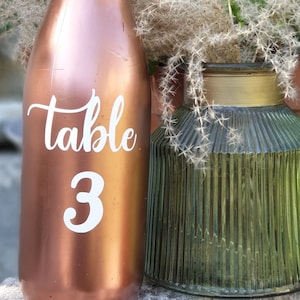 Peut inclure: Une bouteille de couleur cuivre avec une étiquette blanche sur laquelle est inscrit "table 3". La bouteille est à côté d'un vase en verre vert avec un bord doré.