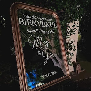 May include: A rectangular mirror with a bronze-colored frame featuring white text that reads "kính chào quý khách", "BIENVENUE", "أهلا وسهلا بالجميع", "Mey & Yoan", and "19 MAI 2026". The mirror reflects the surrounding trees and foliage in a dark setting.