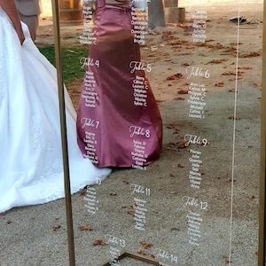 Peut inclure: Un plan de table de mariage en acrylique transparent avec un cadre en métal doré. Le tableau affiche les noms des invités organisés par numéro de table. Un motif floral est dans le coin inférieur droit.