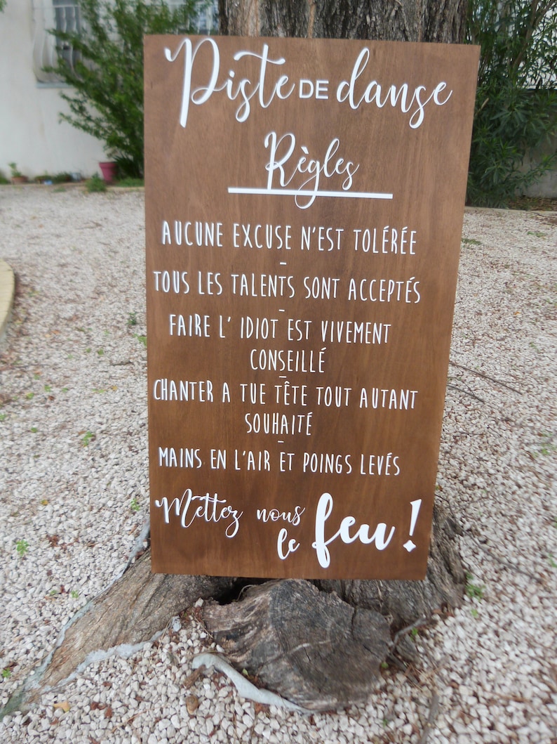 May include: A wooden sign with white text that reads "Piste de danse" and "R&egrave;gles". The sign lists rules for a dance floor, including "AUCUNE EXCUSE N'EST TOL&Eacute;R&Eacute;E", "TOUS LES TALENTS SONT ACCEPT&Eacute;S", "FAIRE L' IDIOT EST VIVEMENT CONSEILL&Eacute;", "CHANTER A TUE T&Ecirc;TE TOUT AUTANT SOUHAITE", "MAINS EN L'AIR ET POINGS LEV&Eacute;S", and "Mettoy nous le feu!"