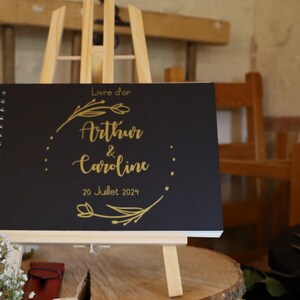 May include: A black spiral-bound guest book with gold lettering that reads Livre d'or, Arthur & Caroline, and 20 Juillet 2024, framed by a gold floral wreath design. The book is displayed on a light-colored wooden easel stand.