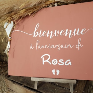 May include: A rectangular terracotta-colored sign featuring white cursive text that reads bienvenue à l'anniversaire de Rosa, accompanied by a small white heart ornament with a tassel and a pair of white baby footprint icons at the bottom.