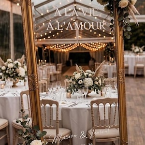 Peut inclure: Un grand miroir doré orné avec le texte "À LA VIE, À L'AMOUR" est installé lors d'une réception de mariage. Le miroir reflète les tables et les chaises décorées. Les noms "Marion & Laurent" et la date "02. 05. 26" sont également affichés.