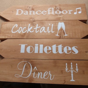May include: Four light brown wooden arrow-shaped signs with white lettering and graphics. The signs read Dancefloor with a music note, Cocktail with two glasses, Toilettes, and Dîner with a candelabra. Each sign features a pointed end.
