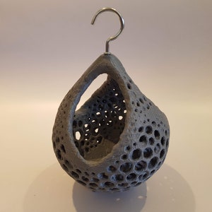 May include: A grey ceramic hanging planter with a unique, honeycomb-like design. The planter has a metal hook at the top for hanging.