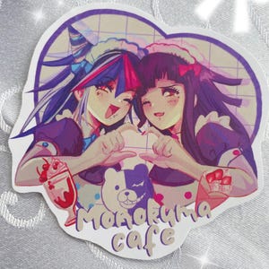 Puede incluir: Una pegatina en forma de corazón con dos personajes de estilo anime con trajes de doncella. Los personajes sonríen y forman un corazón con las manos. La pegatina tiene el texto "Monokuma cafe" y un oso de dibujos animados.