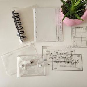 May include: Clear plastic binder, pouches, and cash envelopes. The binder has a black spine. The cash envelopes have snap closures. There are also clear cards with text that reads "One Thousand Dollars". A pink flamingo planter with a succulent is in the background.