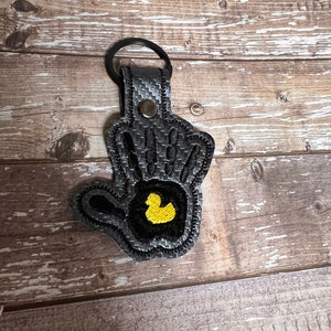 May include: A black and gray key chain with a rubber ducky design. The key chain is made of a textured fabric and has a black metal ring.