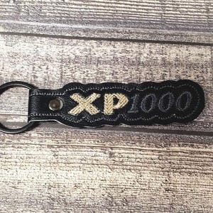 May include: Black leather keychain with a gold embroidered "XP1000" design.
