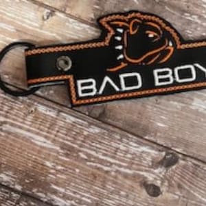 May include: Black keychain with the words "BAD BOY" in white block letters. The keychain features an orange dinosaur graphic and trim. A black key ring is attached to the top left corner. The background is a wooden surface.