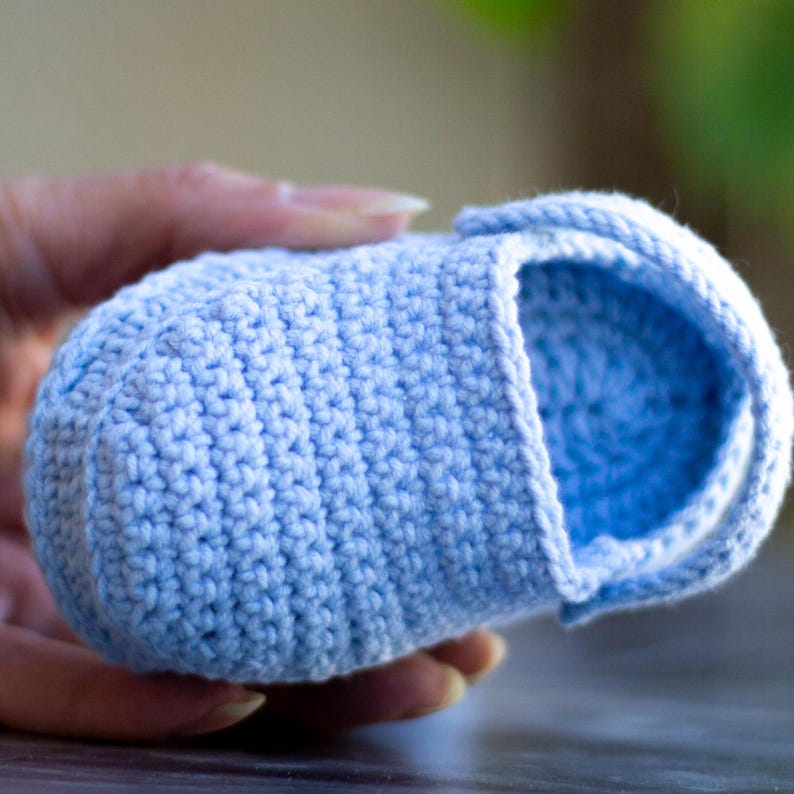 Op de afbeelding: Een lichtblauwe gehaakte babyschoen met een bandje. De schoen is gemaakt met een vaste steek.