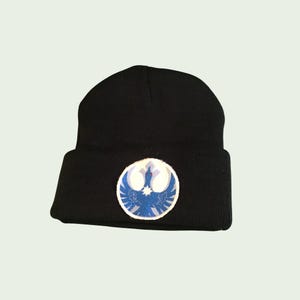 Op de afbeelding: Zwarte beanie-muts met een cirkelvormige patch met een blauw en wit embleem. Het embleem heeft een ster in het midden en een vogelachtig ontwerp. De muts is gemaakt van zacht, gebreid materiaal.