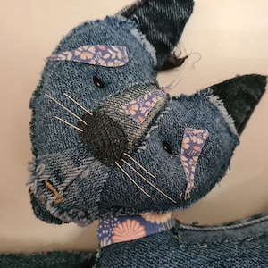 May include: Close-up of a blue denim cat toy with a floral patterned collar. The cat's eyes are black buttons and its whiskers are made of white thread.