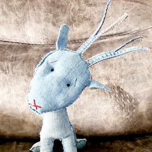 Peut inclure: Un jouet animal en denim bleu avec un long cou et des bois. Le jouet a un visage cousu avec des yeux noirs et une bouche rouge.