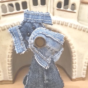 Può includere: Un piccolo giocattolo di jeans a forma di cane, in piedi su una superficie di legno davanti a una casa di ceramica bianca. Il giocattolo ha un bottone con il testo "Quality Product" su di esso.