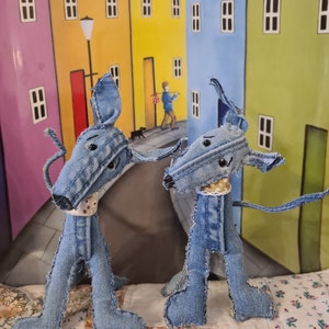 Può includere: Due cani giocattolo in denim blu in piedi su una trapunta a motivi. I cani hanno gli occhi marroni e sono fatti di tessuto di denim riutilizzato. I cani sono in piedi davanti a un dipinto di una fila di case colorate.