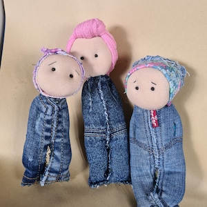 May include: Three handmade fabric dolls with denim jean jackets and fabric heads. The dolls have pink, blue, and purple head coverings. The dolls have black button eyes and stitched mouths. The dolls are on a tan background.