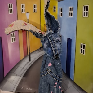 May include: A blue denim bunny rabbit with a white fabric ear and pink embroidered flowers. The bunny is standing in front of a colorful painting of houses.