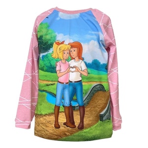 Könnte beinhalten: Ein langärmliges Shirt mit einer Cartoon-Illustration von zwei Mädchen, die mit ihren Händen ein Herz formen. Das Shirt hat rosa Ärmel mit einem weißen geometrischen Muster. Der Hintergrund der Illustration zeigt eine Brücke, einen Bach und einen blauen Himmel.