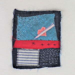 Op de afbeelding: Een klein, ingelijst stuk textielkunst met een blauwe achtergrond, een rood hart en een witte lijn. De lijst is gemaakt van zwarte stof en heeft een rode streep met de symbolen 'x' en '+' erop.