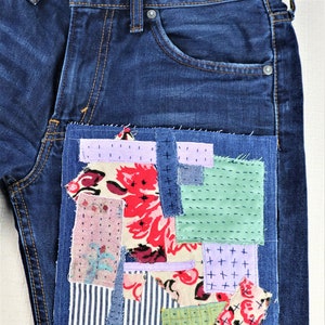 以下が含まれることがあります： 前面のポケットにパッチワークのデザインが施された青いデニムジーンズ。パッチワークは、花柄、ストライプ、チェックなど、さまざまな色のさまざまな生地で構成されています。