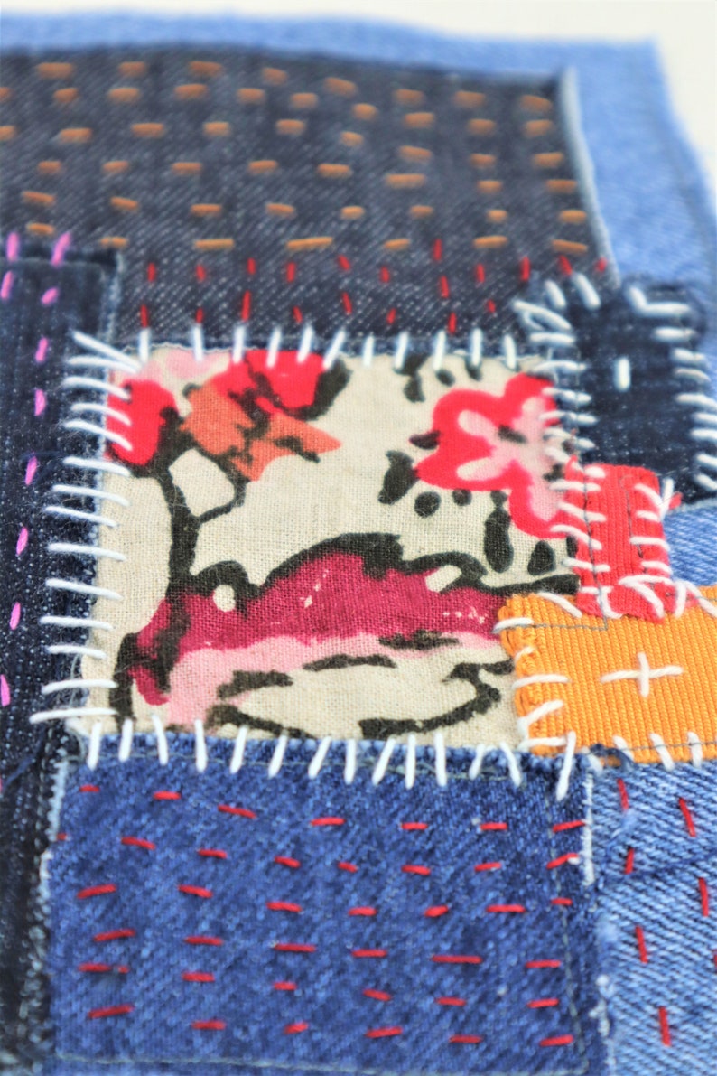 May include: A patchwork of denim fabric in shades of blue, with a floral print fabric in the centre. The fabric is stitched together with white thread, and there are red and orange accents.