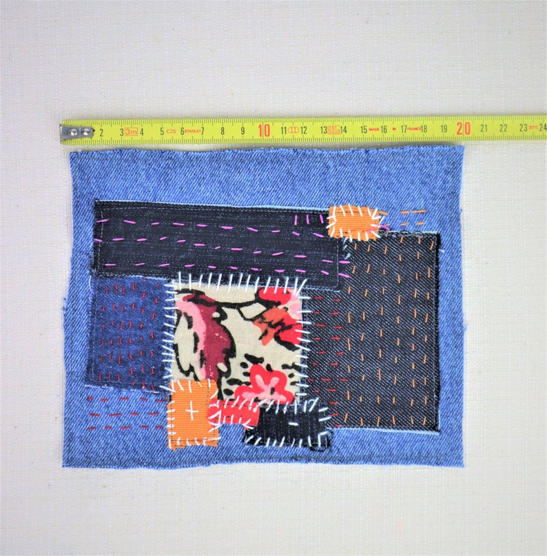 May include: A blue denim patchwork quilt with various fabric scraps sewn together. The quilt features a floral print fabric, a solid orange fabric, and a black denim fabric. The quilt is stitched with white, pink, and orange thread.