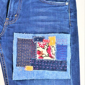 May include: Blue denim jeans with a patchwork design on the front pocket. The patchwork is made of different coloured denim fabrics and includes a floral print.