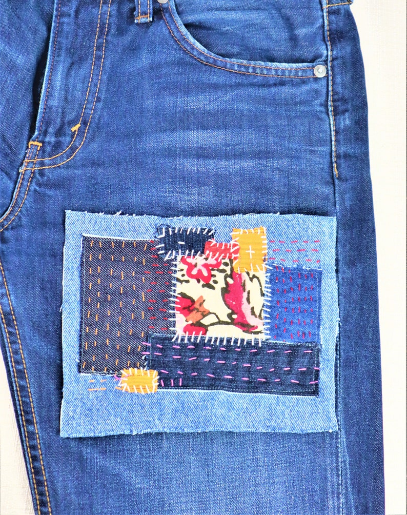 May include: Blue denim jeans with a patchwork design on the front pocket. The patchwork is made of different coloured denim fabrics and includes a floral print.