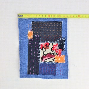 May include: A blue denim patchwork square with different coloured fabrics sewn together. The fabrics include a floral print, solid orange, and black denim. The square is surrounded by a blue denim border.
