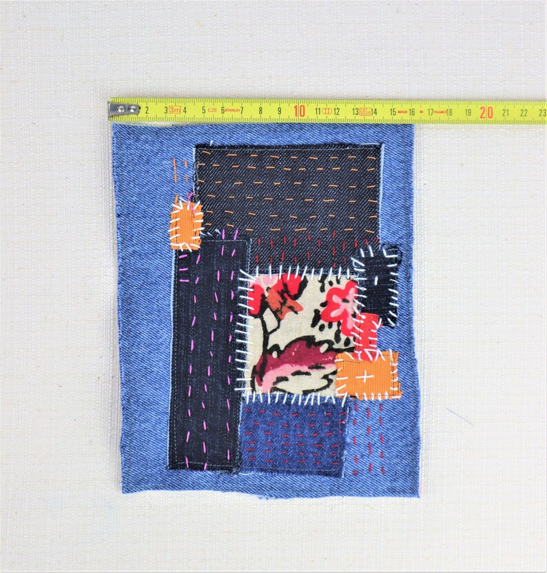 May include: A blue denim patchwork square with different coloured fabrics sewn together. The fabrics include a floral print, solid orange, and black denim. The square is surrounded by a blue denim border.